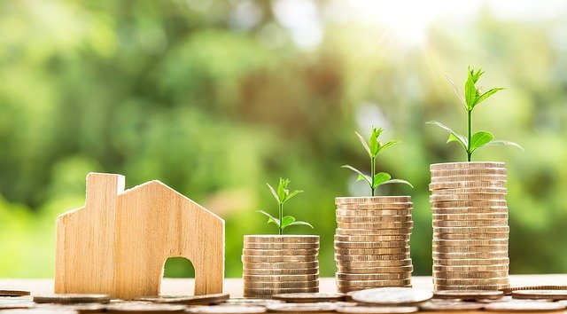 Pequeña casa ecológica junto a montones de dinero con plantas creciendo en la parte superior.